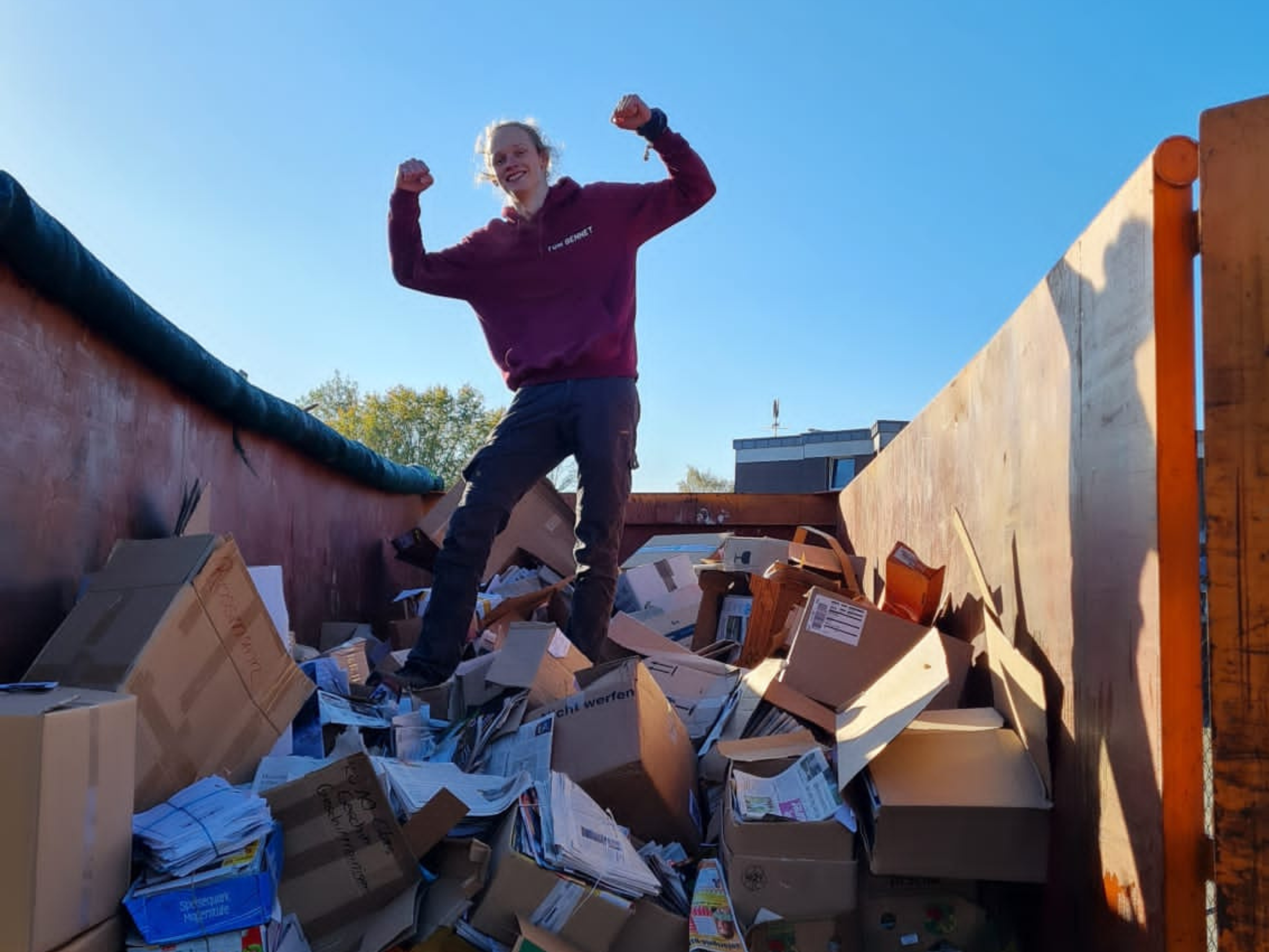 Person steht mit erhobenen Armen auf einem großen Haufen aus zerknittertem Altpapier und Kartons in einem Container bei der Altpapiersammlung